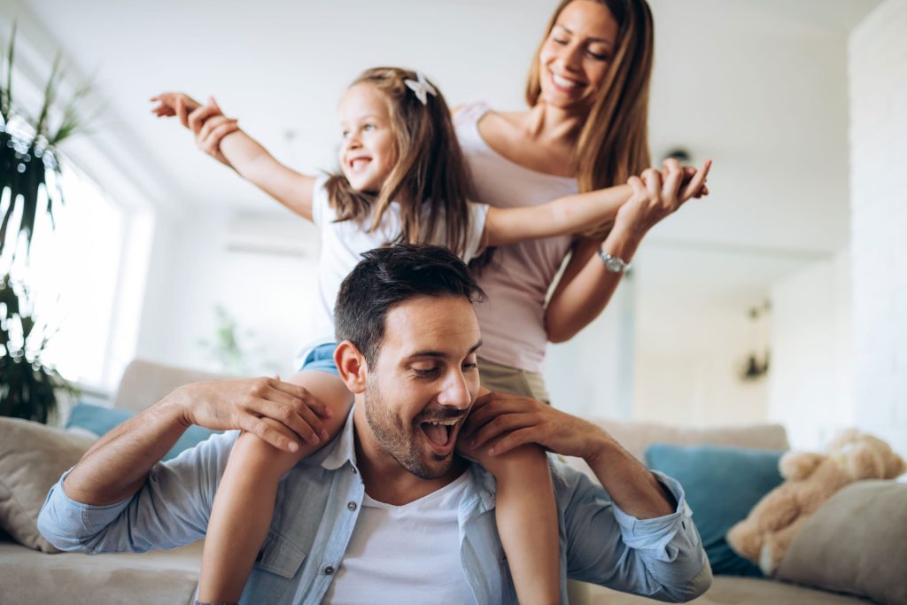 Happy family having fun at home with a young child on her fathers shoulders with outstretched arms and a mother holding the child in the background